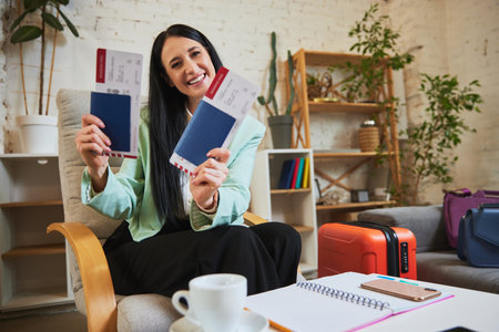 Happy smiling businesswoman sitting at home and showing passport with tickets, expressing excitement about upcoming vacationの写真素材