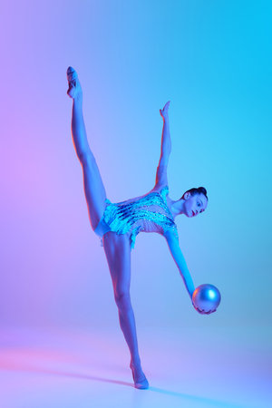 Full-length dynamic image of young talented girl in sparkling costume performing with ball against gradient studio background in neon lightの写真素材