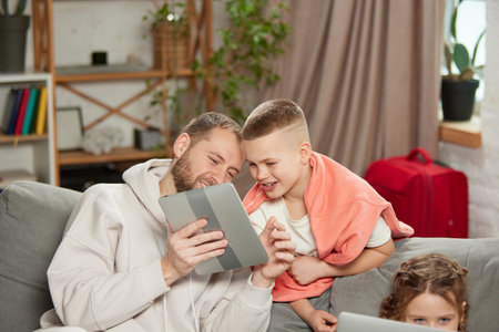 Photo of father with son, man with boy looking at tablet and smiling while little girl sitting with laptop.の写真素材