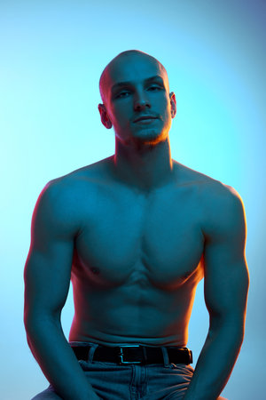 Portrait of handsome bald young man with muscular, shirtless, sportive body sitting in jeans against blue studio background in neon lightの写真素材