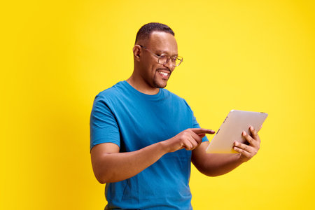 Smiling African-American man in glasses and blue t-shirt using tablet for online shopping services...