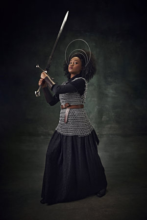 Brave young African woman, medieval warrior in chainmail armor with halo-like rings above head making sword swing against vintage backgroundの写真素材