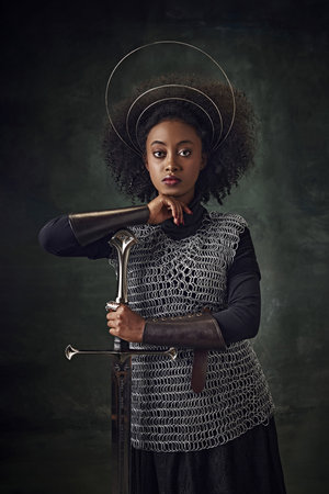 Brave young African woman, medieval warrior in chainmail armor with halo-like rings above head leaning sword on dark green vintage backgroundの写真素材