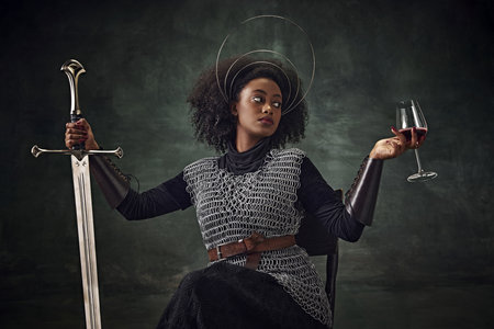 African woman, medieval warrior in chainmail armor sitting on chair with sword and wine glass against vintage green background. Brave womanの写真素材