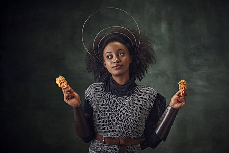Young African woman, medieval warrior in chainmail armor holding fried chicken against vintage green...