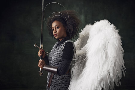 Young African woman, medieval warrior with angel wings, halo above head and chainmail holding sword against vintage dark backgroundの写真素材