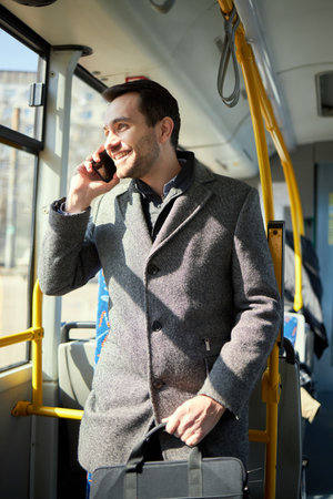 Man in grey coat standing in modern tram, talking on mobile phone wit smile. Fast and convenient way of transportationの写真素材