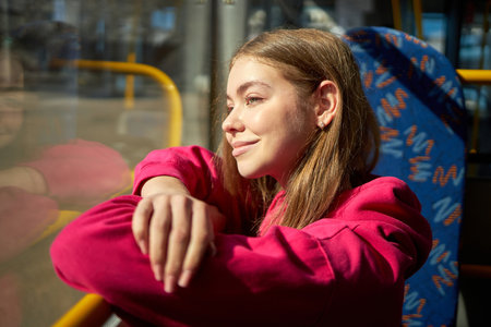 Young girl in pink hoodie sitting in public transport, modern tram, bus, looking in window and...