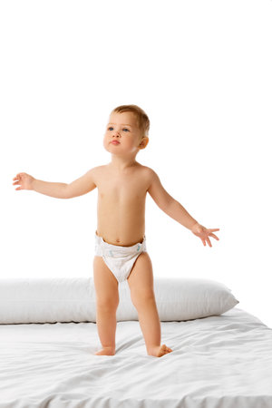 Adorable little child, baby boy standing on bed in diaper and curiously looking isolated over white studio backgroundの写真素材