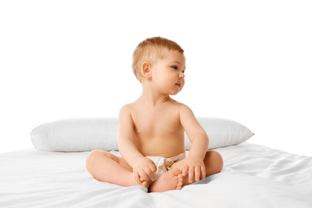 Beautiful little baby, boy, child in diaper calmly sitting on bed isolated over white studio backgroundの写真素材