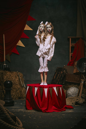 Siamese twin girls in white vintage costumes with makeup stand on stage over dark retro circus...