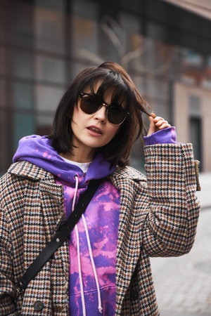 Portrait of young brunette woman in stylish sunglasses, colorful hoodie and classic coat posing against urban backgroundの写真素材