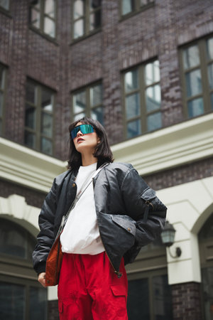 Contemporary urban clothing outlets. Bottom view photo of elegant, beautiful young woman in red cargo pants and stylish bomber jacketの写真素材