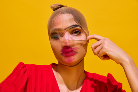 Young woman with stocking over head with smudged lipstick makeup against yellow background....