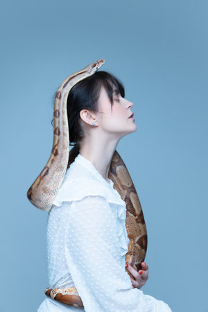 One direction way. Portrait of brunette young woman in white dress sitting with closed eyes and snake on her head against blue studio background.の写真素材