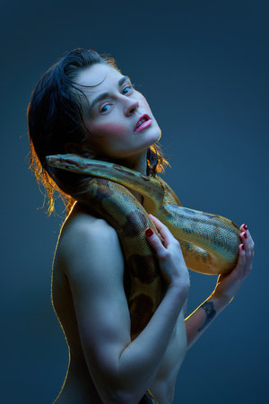 Portrait of young elegant woman with pink cheeks posing shirtless with snake over her body against...