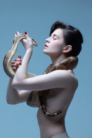 Close to nature. Beautiful brunette young woman with slim body posing with snake on blue studio background.の写真素材