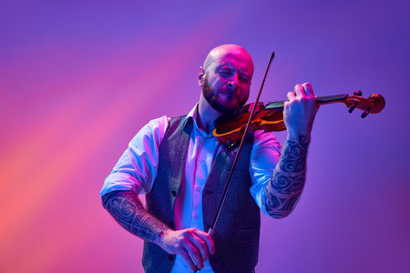 Portrait of talented young man, solo performer playing violin against gradient purple background in neon lightの写真素材