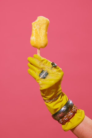 Female hand in rubber cleaning yellow gloves with jewelry, accessories holding melting ice cream with soap bubbles against pink background.の写真素材