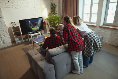 Emotional young people, friends gathering in living room at home, eating snacks, drinking beer and watching online football match translationの写真素材