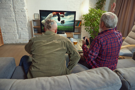 Two elderly men, sport fans sitting on couch at home in living room and watching online football match translation on tv. Supporting teamの写真素材