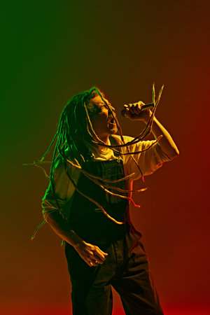 Young man, passionate musician with dreadlocks emotionally singing against gradient red green background in neon light.の写真素材