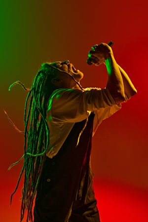 Emotive man, soulful musician with dreadlocks expressing his deepest emotions through his singing against gradient red green background in neon lightの写真素材