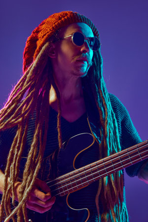 Young man with dreadlocks, beanie hat and sunglasses creating beautiful music against purple background in neon lightの写真素材