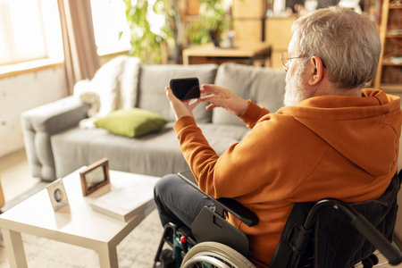Elderly man sitting on wheelchair at home in living room and watching news, videos on mobile phone....