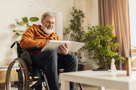 Senior positive man sitting on wheelchair at home in cozy light living room and reading book. Leisure time, relaxationの写真素材