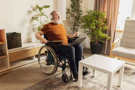 Elderly man sitting on wheelchair at home in living room in headphones and learning how to play...