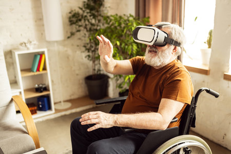 Elderly man sitting on wheelchair in living room at home and wearing VR glasses. Exploring innovative technologiesの写真素材