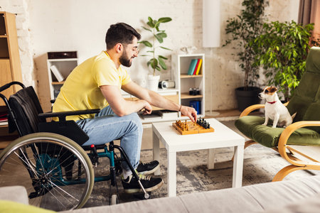 Smiling young man sitting on wheelchair in living room at home and playing chess with his little dog. Leisure timeの写真素材