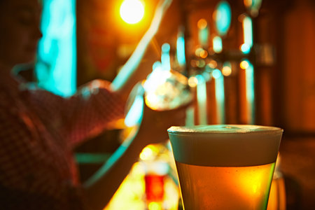 Focus on glass with foamy lager beer and blurred image of bartender pouring another glass of beer in...