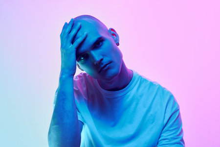 Portrait of young man with bald head, shaved face, inn casual t-shirt posing on gradient blue pink background in neon lightの写真素材