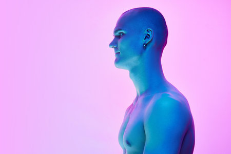 Side view image of young bald man with shirtless muscular body posing on gradient blue pink background in neon lightの写真素材