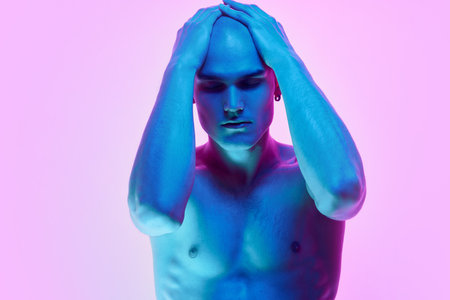 Self-identity. Portrait of young man with bald head, posing shirtless on gradient blue pink background in neon lightの写真素材