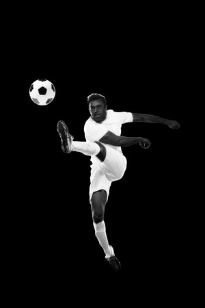 Monochrome dynamic image of young man, soccer player in motion hitting ball in a jump on black backgroundの写真素材
