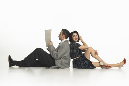 Happy young woman in retro-style dress sitting back to back with man in suit reading newspaper...