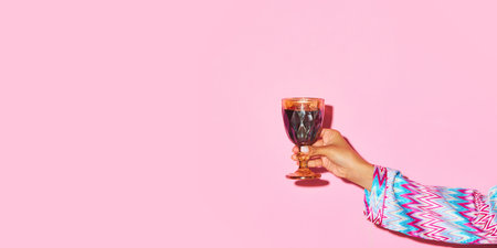 Female hand holding glass with red wine against pink background. Toast, celebration.の写真素材