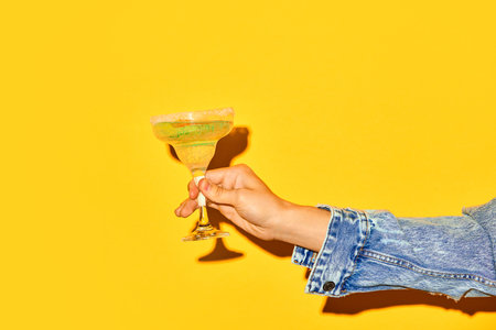 Male hand holding sweet and sour margarita cocktail with slice of orange against bright yellow backgroundの写真素材