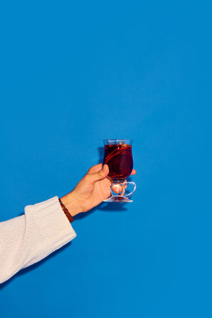 Hand holding glass with mulled wine made with red wine and cinnamon against blue background. Traditional winter drinkの写真素材