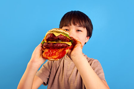 Little boy, kid holding giant, juicy fresh burger with meat, tomato sauce and cheese, eating with delightful face against blue backgroundの写真素材