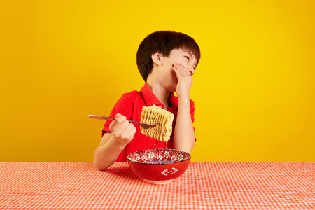 Cheerful child in red polo shirt sitting at table, laughing and eating instant noodles with fork against yellow backgroundの写真素材