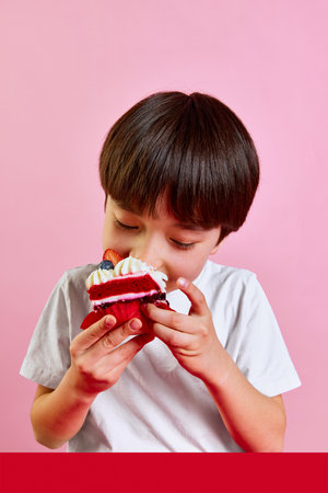 Delightful dessert time. Boy, kid enjoying delicious red velvet cake with cream and berries, making big bite against pink backgroundの写真素材