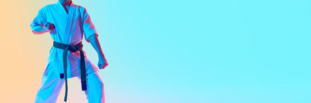 Cropped image of boy, karate athlete in uniform, green belt standing against gradient orange blue background in neon lightの写真素材