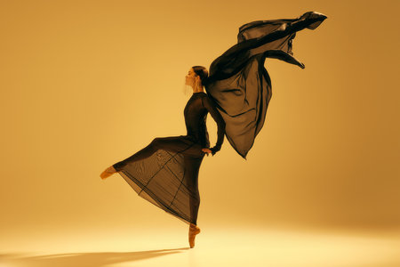 Dynamic ballet movement. Full-length image of artistic female ballet dancer, ballerina in black flowing clothes dancing against yellow backgroundの写真素材