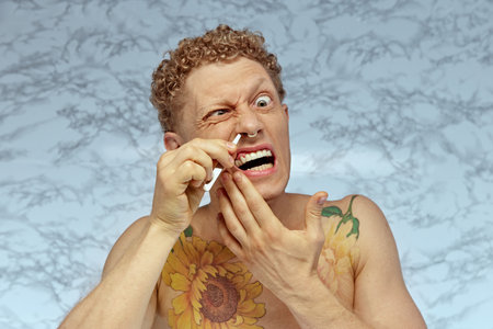 Funny image of shirtless emotional man taking care after his look, using tweezers for nose hair removal. Beauty routineの写真素材
