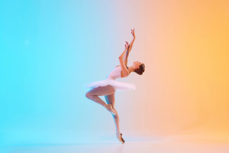 Young ballerina in white tutu dancing and leaning backwards in motion in neon light against blue-orange gradient background.の写真素材