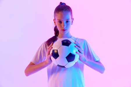 Concentrated football player, teen girl in uniform posing with ball against pink studio background in neon lightの写真素材